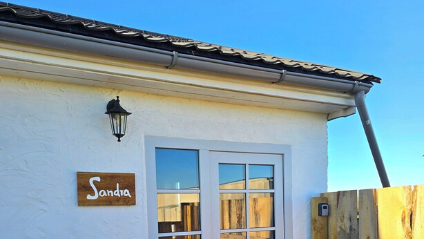 A white building with a wooden nameplate reading "Sandra," a wooden door with glass panels, and a wooden deck. - Vacation apartment "Sandra" in Denmark  (Bredebro)