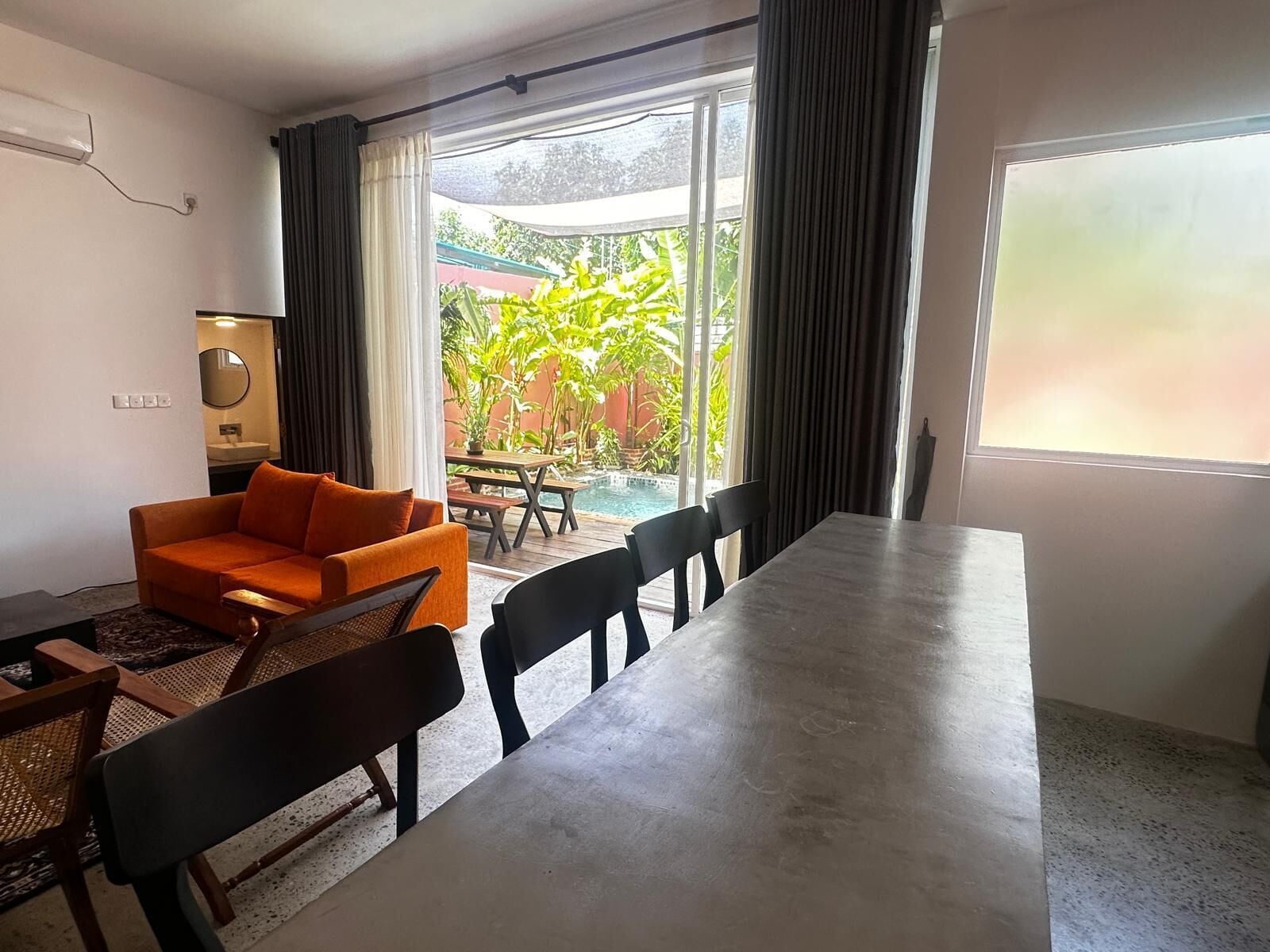 Ein Zimmer mit Esstisch, Stühlen, einer Couch, einer Bank und Blick auf ein Schwimmbad und Grünflächen.