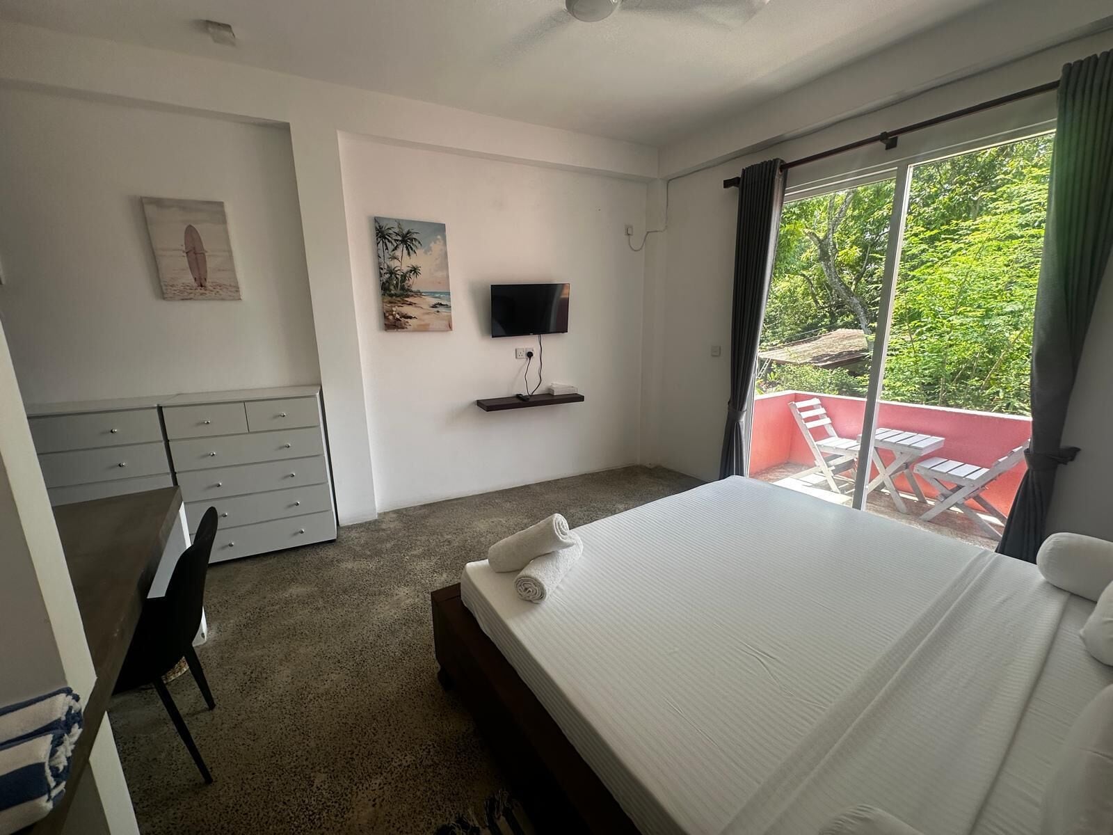 Hotelkamer met een bed, bureau, stoel, televisie en balkon met een rode muur en witte stoelen.