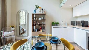 Ein modernes Wohnzimmer mit einem Glastisch, gelben Stühlen, einem Bücherregal und einer Küchenzeile inklusive Mikrowelle und Kaffeemaschine.