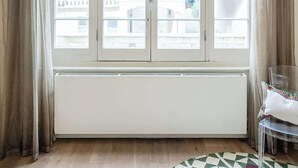 Ein Zimmer mit einem großen Bogenfenster, einem Heizkörper, einem klaren Stuhl und einem gemusterten Teppich.