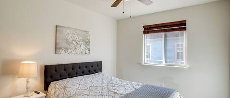 A bedroom with a bed, a nightstand, a lamp, a ceiling fan, a window with blinds, and a wall-mounted artwork.