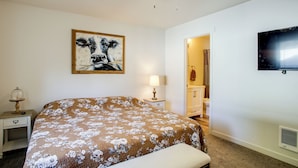 Een slaapkamer met een bed, een plafondventilator, een schilderij, een televisie, en een badkamer zichtbaar door een geopende deur.