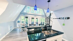 Une cuisine moderne dotée d’un îlot central, de plans de travail noirs et d’un lave-vaisselle. On aperçoit, à travers les fenêtres, un coin repas avec une table et des chaises.