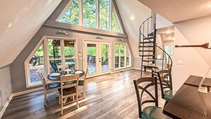 Un loft spacieux avec de hauts plafonds, un coin repas avec une table et des chaises, un escalier en colimaçon et de grandes fenêtres offrant une vue sur l’extérieur.