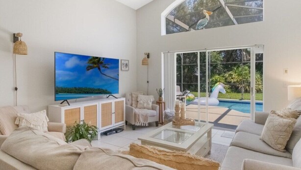 Uma sala de estar espaçosa com uma televisão grande, assentos confortáveis, uma mesa de centro de vidro e vista para uma piscina e um jardim.