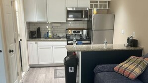 Pieni, käytännöllinen keittiö, jossa on valkoiset kaapit, mikroaaltouuni ja jääkaappi. Työtasolla on pieni roskakori ja vesipullo. Etualalla on sininen sohva, jossa on ruutukuvioinen koristetyyny.