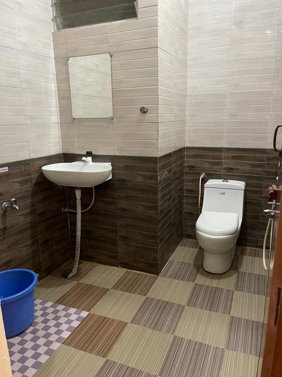 A small bathroom with a sink, toilet, and shower. The floor has a checkered pattern, and there is a blue bucket on the left.