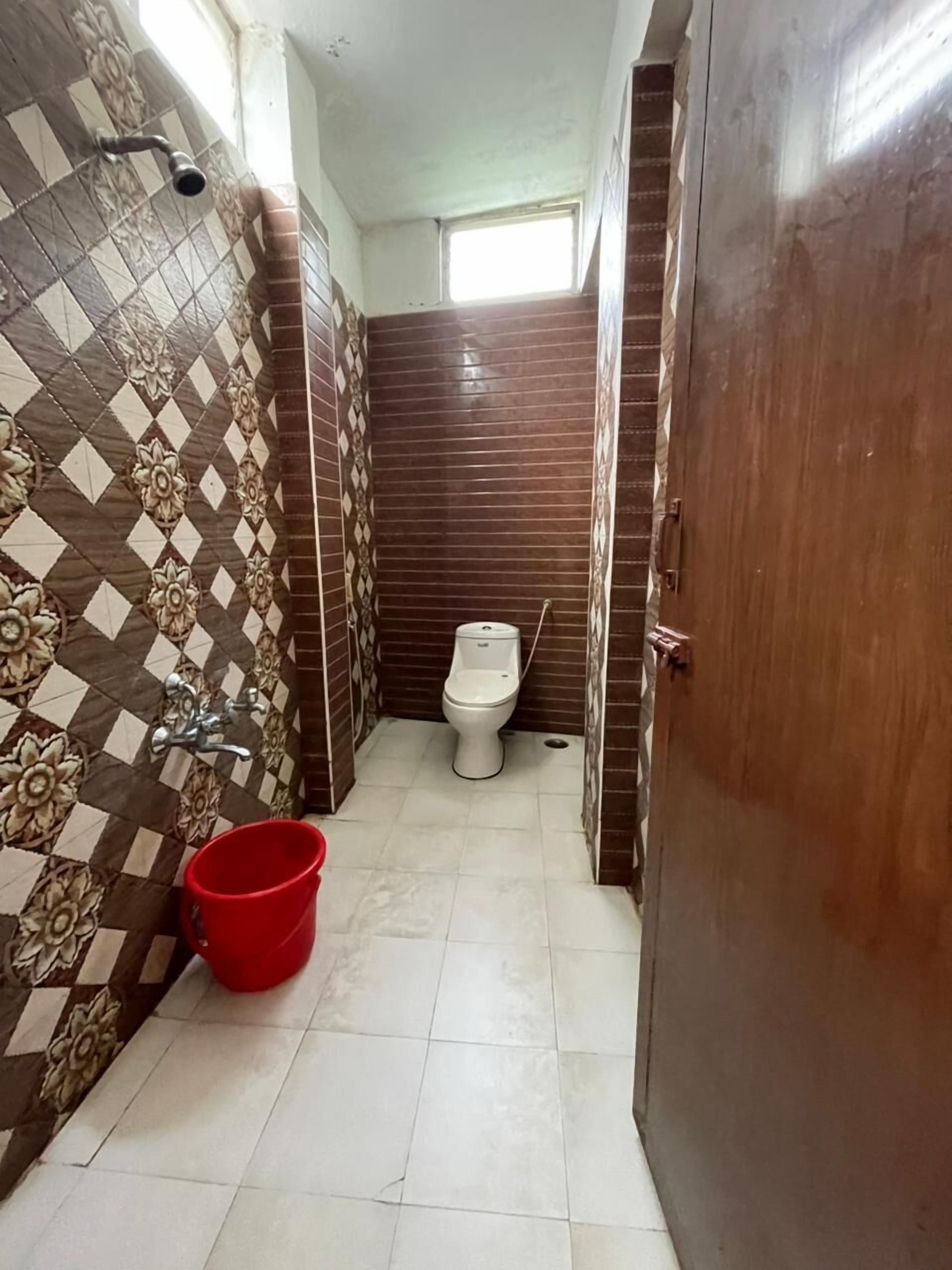 A small bathroom with a toilet, a shower, and a red bucket on the floor.
