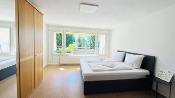 Quarto com cama, guarda-roupa, radiador e janela com vista para o verde.