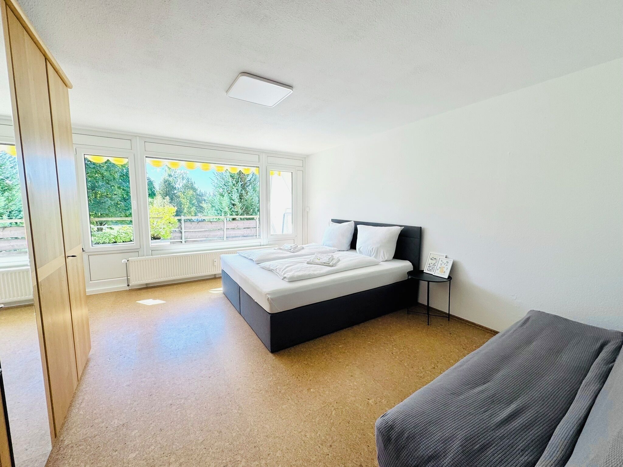 Ein Schlafzimmer mit Bett, Nachttisch, Heizkörper und einem Fenster mit Blick ins Grüne.