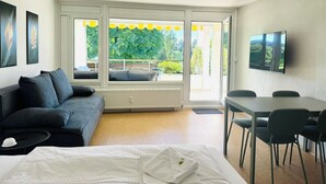 Moderni hotellihuone, jossa on suuri sänky, sohva, ruokapöytä tuoleineen ja seinälle asennettu televisio.
