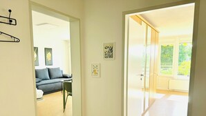Käytävän päästä on pääsy olohuoneeseen, jossa on sohva ja pöytä, ja toisesta ovesta pääsee valoisaan huoneeseen, jossa on ikkuna.