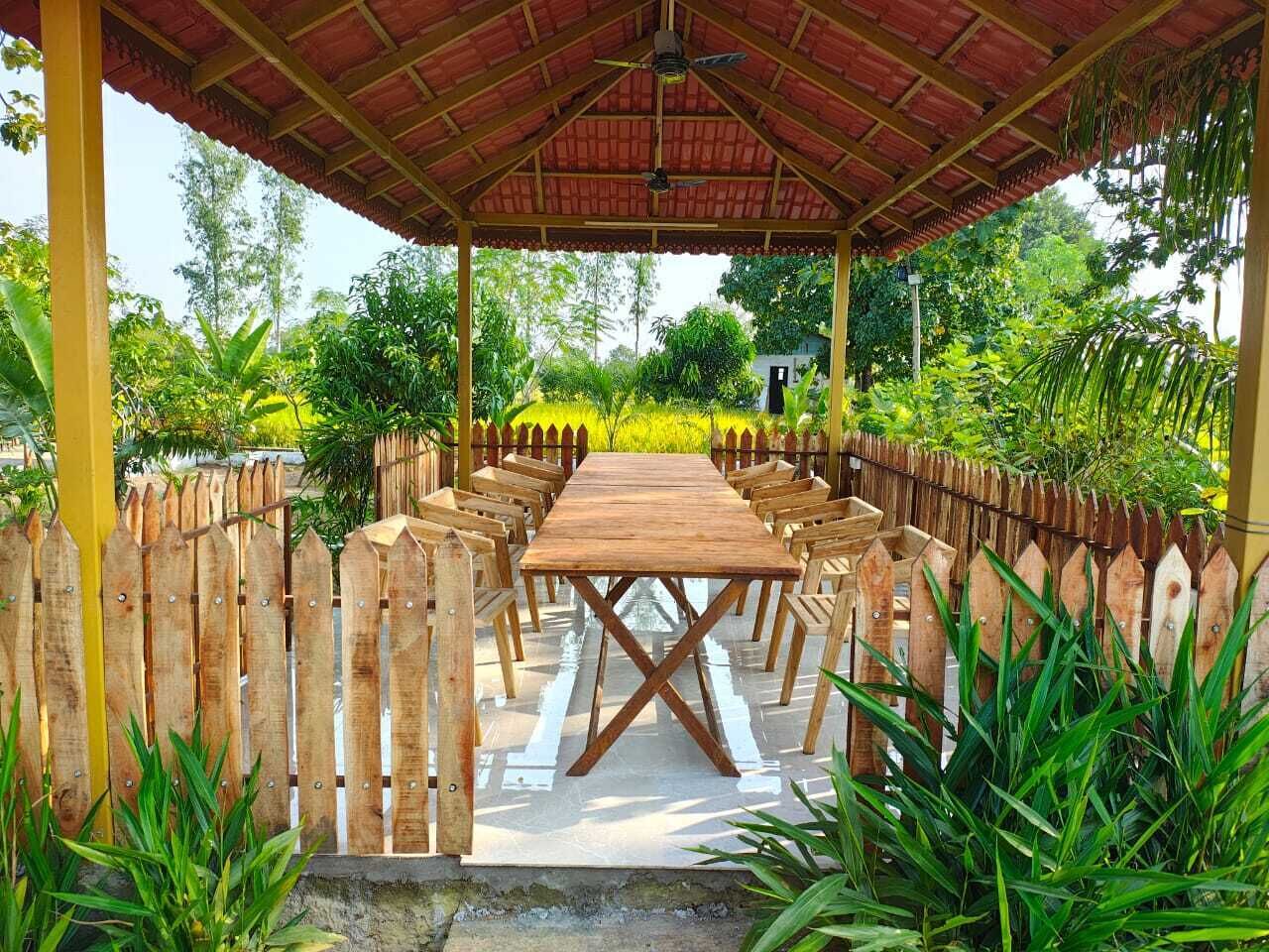 A covered outdoor dining area with a long wooden table and chairs, surrounded by a wooden fence, set in a lush garden.