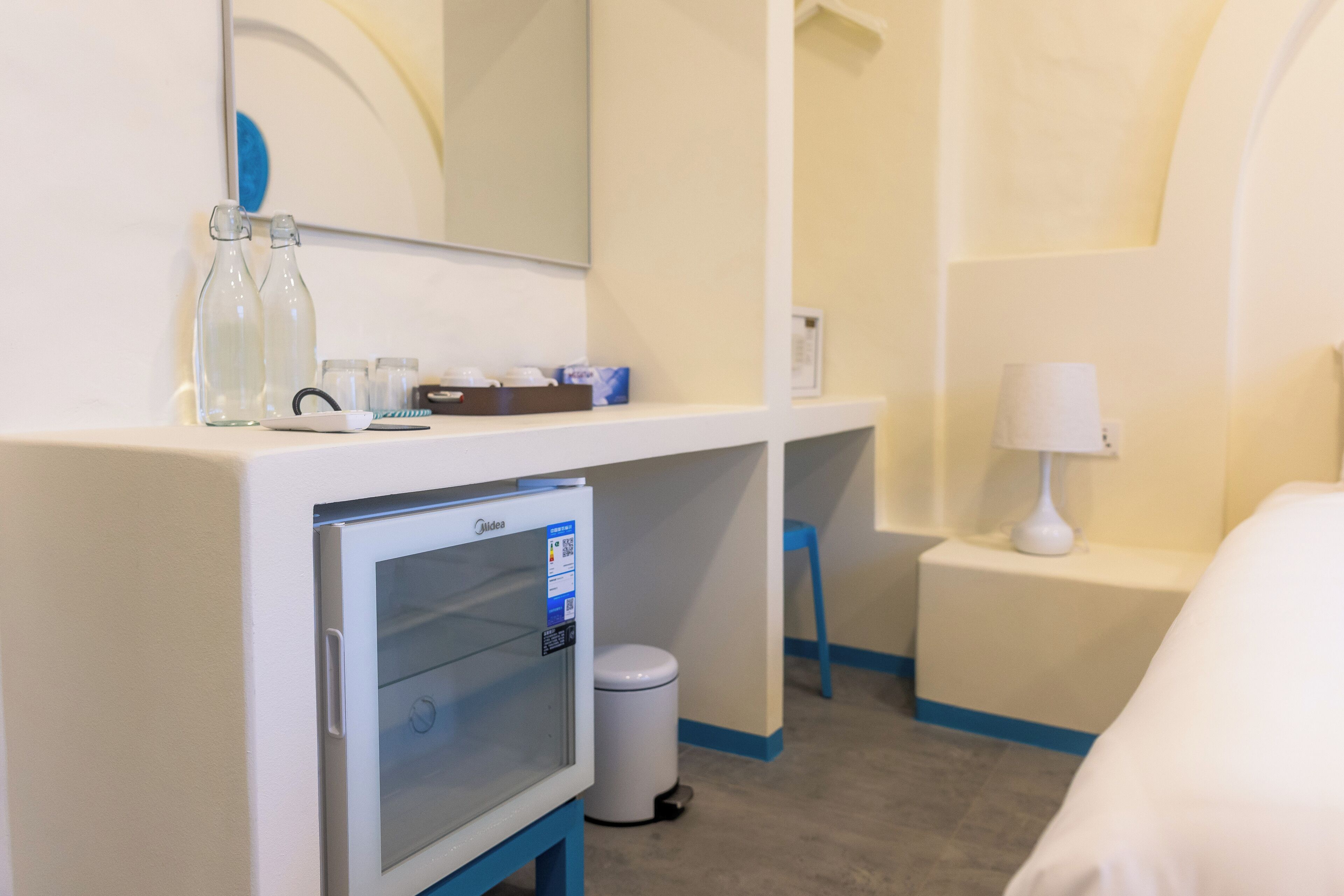 Hotelkamer met een minibar, een spiegel, een kleine prullenbak, een nachtkastje met een lamp en een bed.