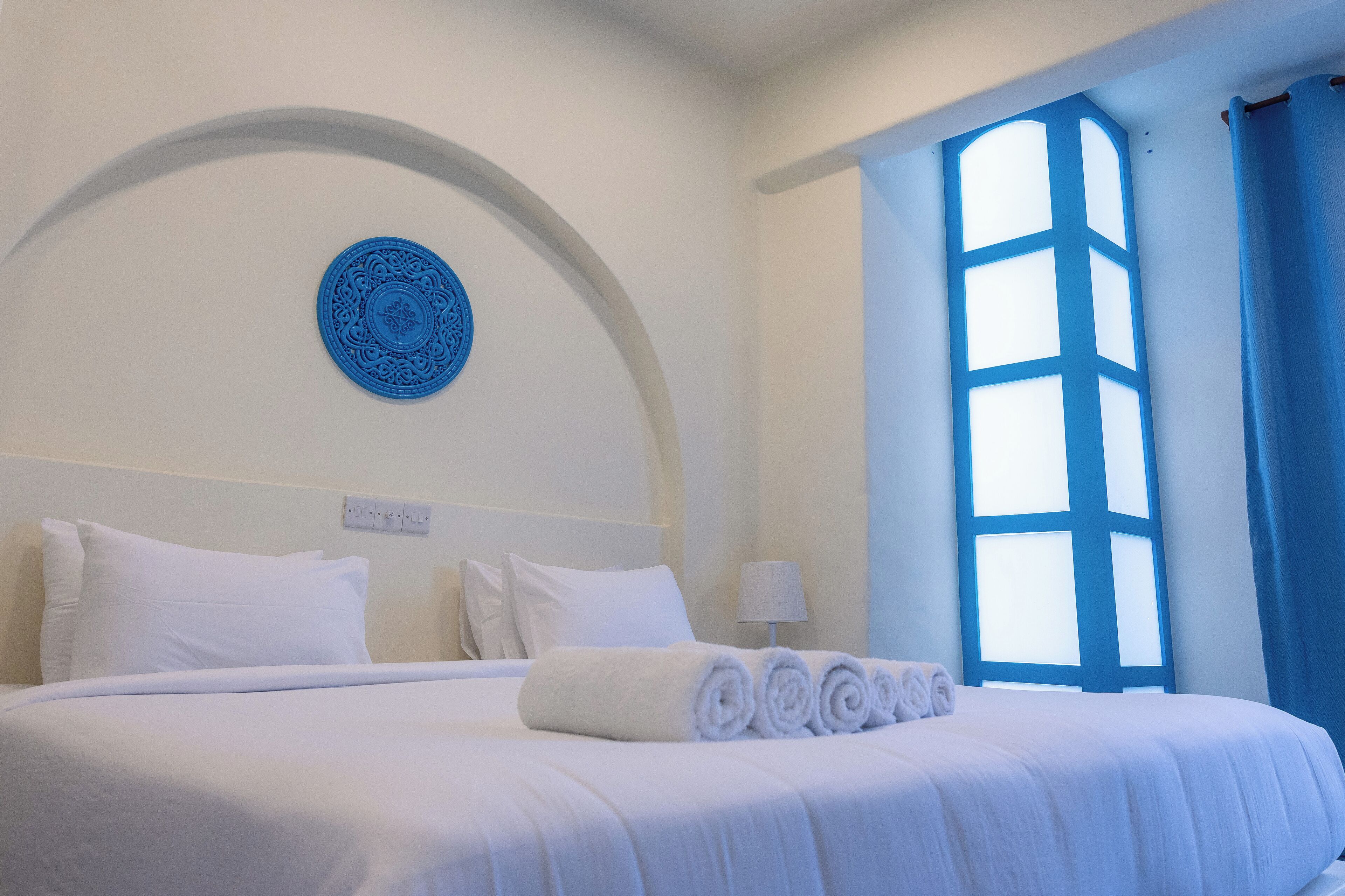 Ein ordentlich gemachtes Bett mit weißen Leinen und aufgerollten Handtüchern, ein blauer dekorativer Teller an der Wand und ein großes Fenster mit blauen Vorhängen.