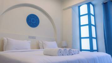 Ein ordentlich gemachtes Bett mit weißen Leinen und aufgerollten Handtüchern, ein blauer dekorativer Teller an der Wand und ein großes Fenster mit blauen Vorhängen.