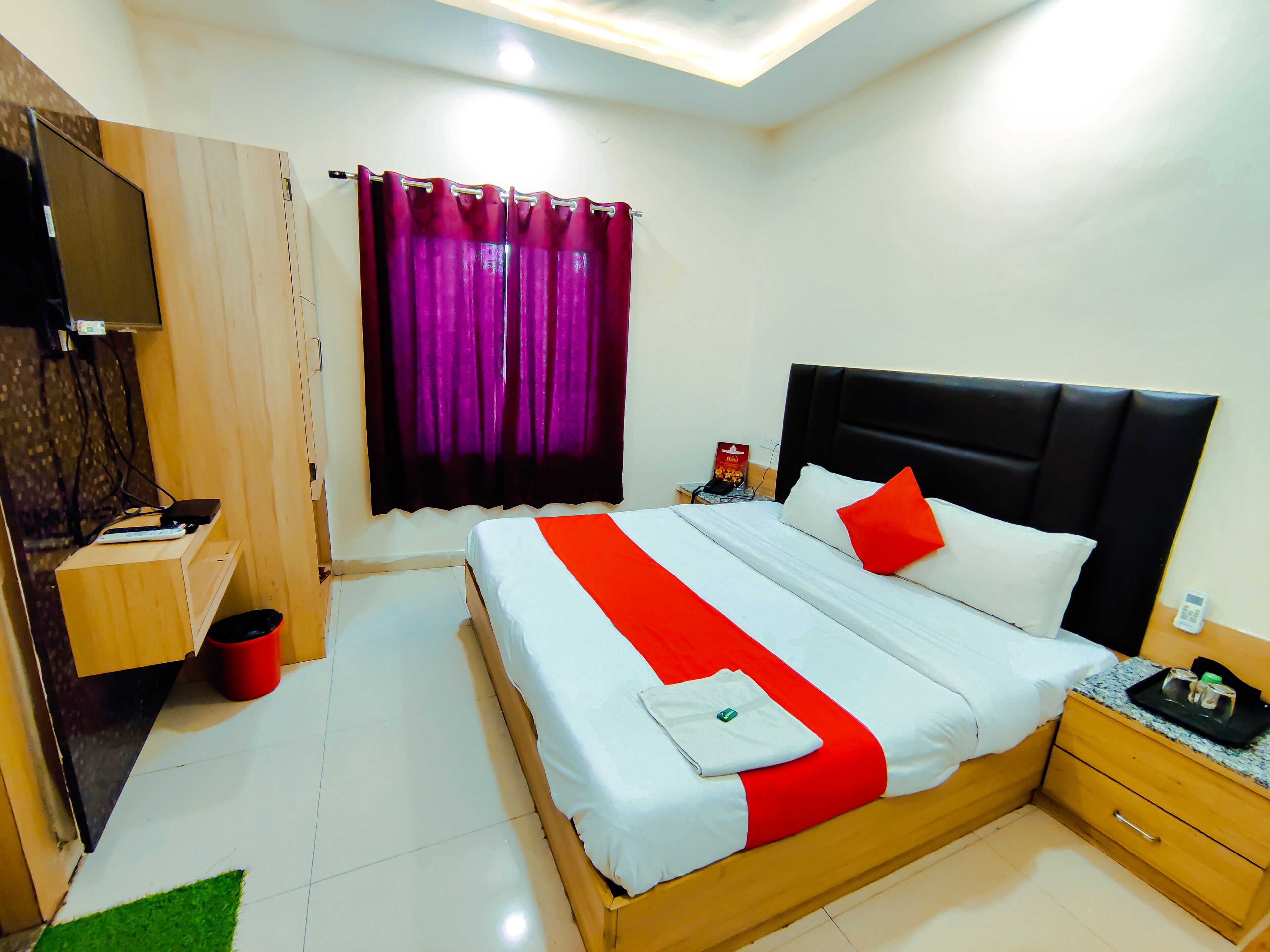 A hotel room with a bed, a nightstand, a desk, a red trash bin, and a window with purple curtains.