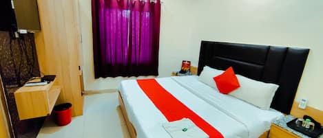 A hotel room with a bed, a nightstand, a desk, a red trash bin, and a window with purple curtains.