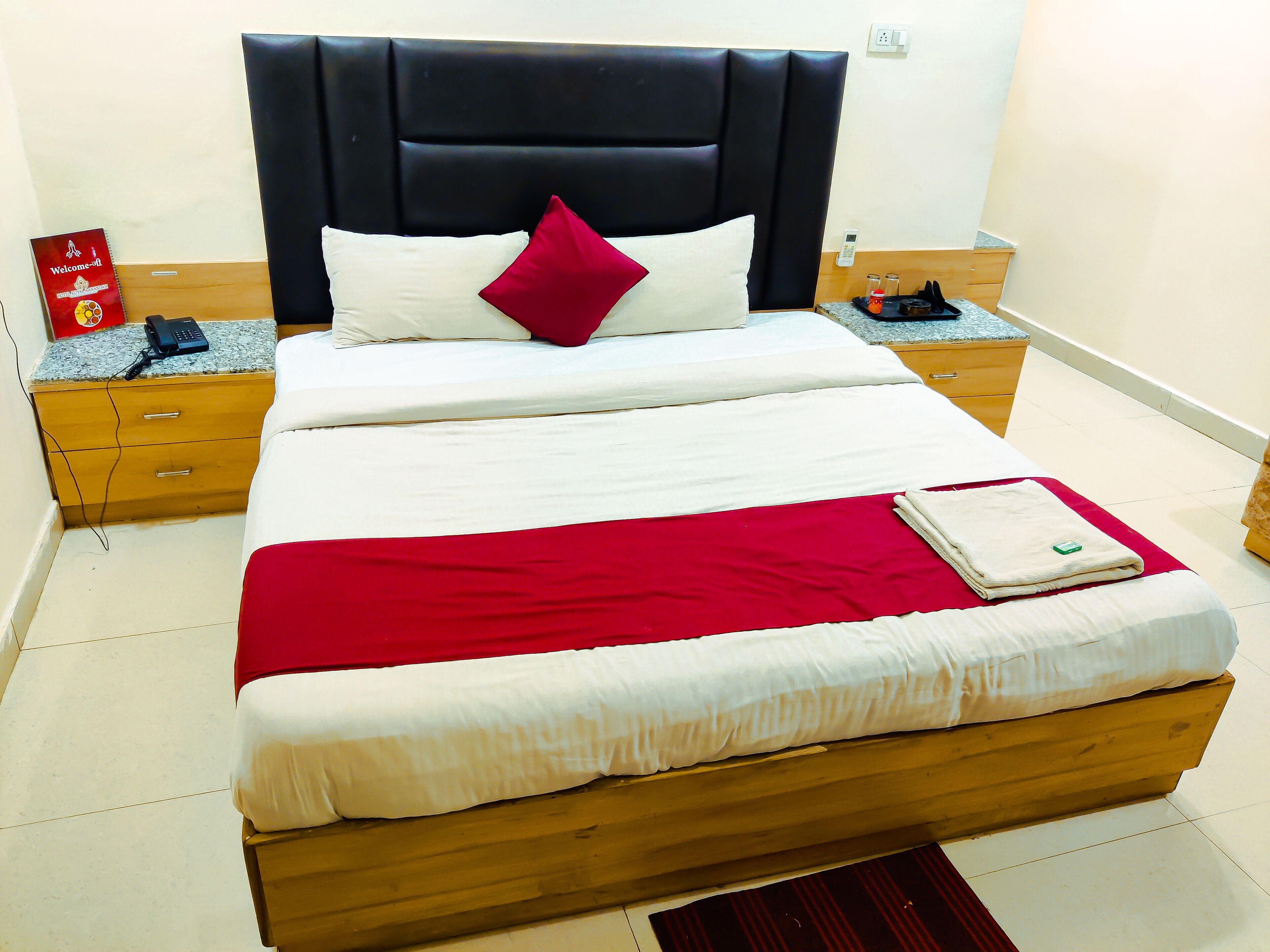 Un lit bien fait, recouvert d’une couverture rouge et de draps blancs, une table de chevet en bois avec un téléphone et un panneau rouge, et une commode en bois avec des tiroirs.