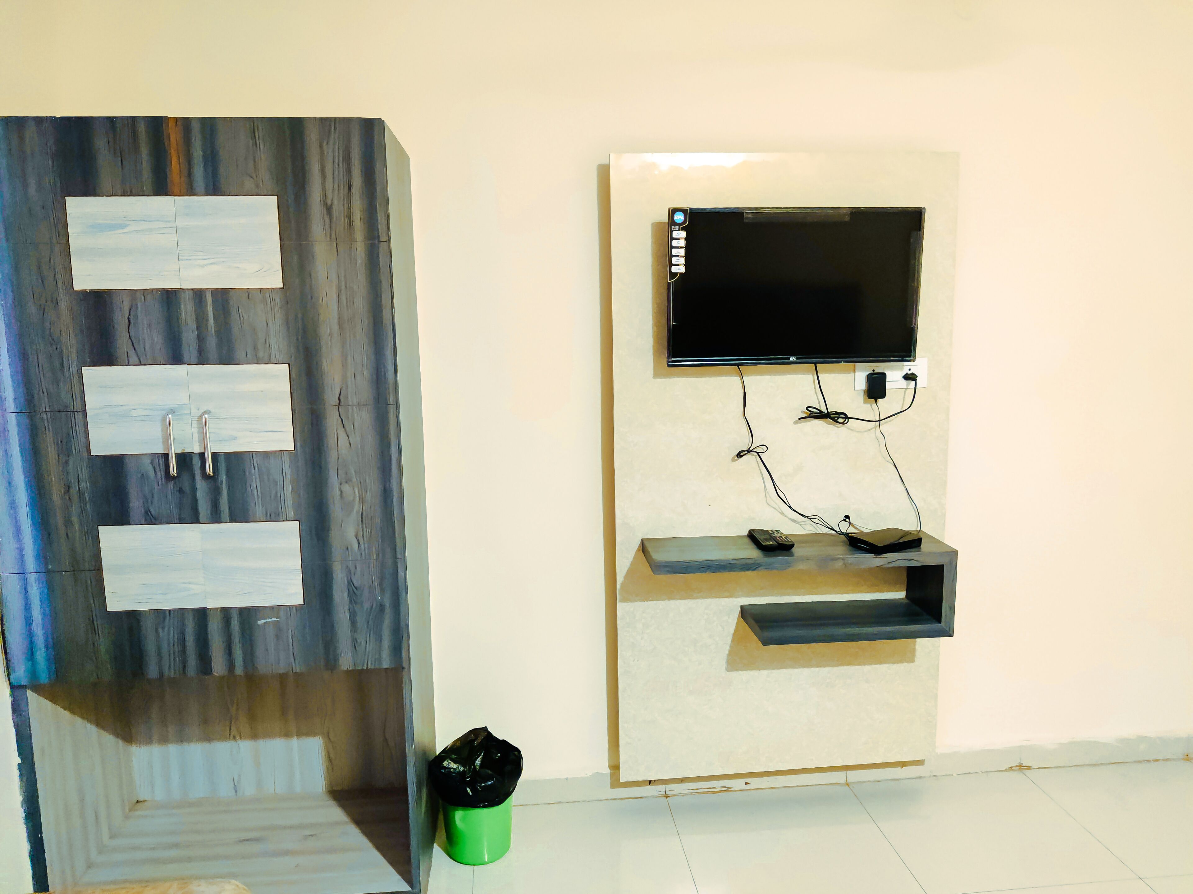 Un televisor de pared con control remoto y una regleta, un cesto de basura verde y un mueble de madera con cajones.