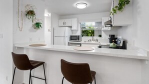 Una cucina moderna con mobili bianchi, un forno a microonde, un frigorifero e una macchina per il caffè. Sono presenti due sgabelli da bar e una pianta pensile.