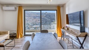 A modern living room with a large sliding glass door leading to a balcony, a flat-screen TV mounted on the wall, and a dining table with chairs. - Stylish Apartment - Breathtaking Views in Xlendi (Munxar)