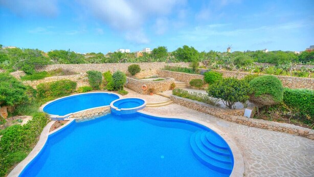 Uma grande piscina ao ar livre com um caminho de pedra que a leva, cercada por vegetação exuberante e um muro de pedra.