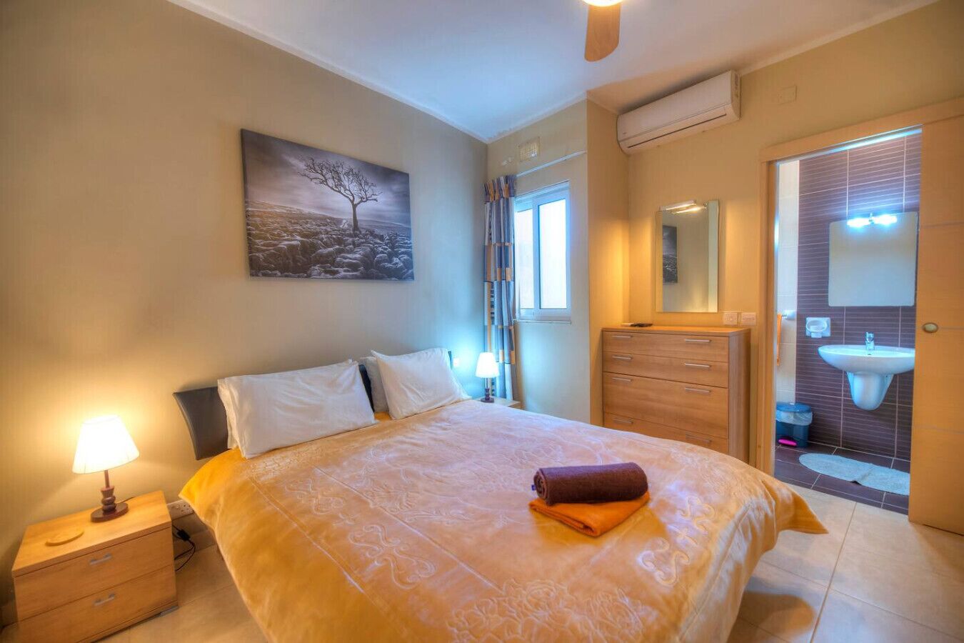 Una camera da letto con un letto, comodino, lampada, cassettiera e bagno con lavandino e specchio.
