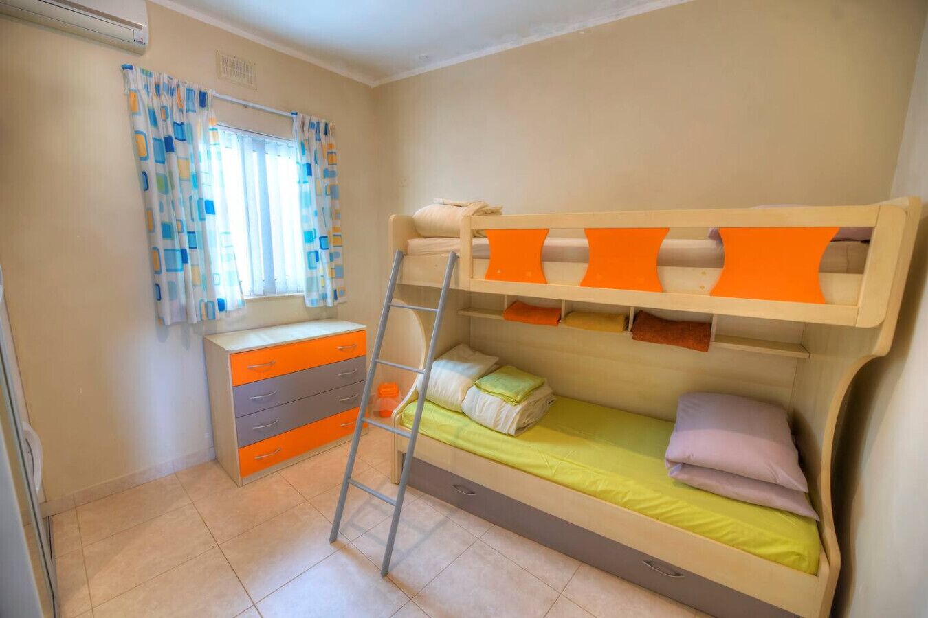 A bunk bed room with a desk and drawers, a ladder, and a window with curtains.