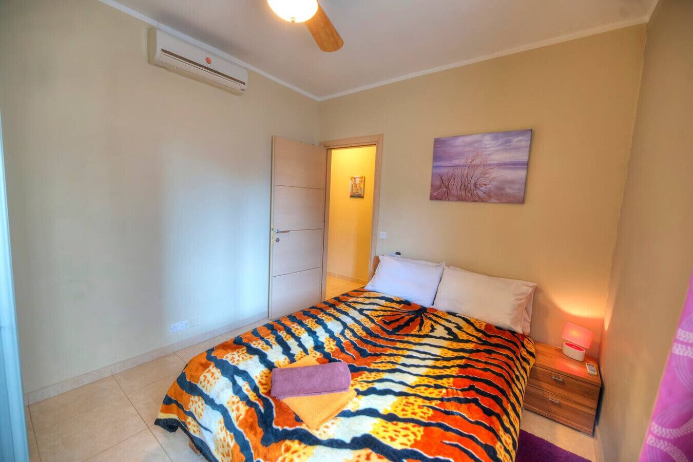 Un dormitorio con cama, una mesita de noche, aire acondicionado de pared, una puerta y un cuadro en la pared.
