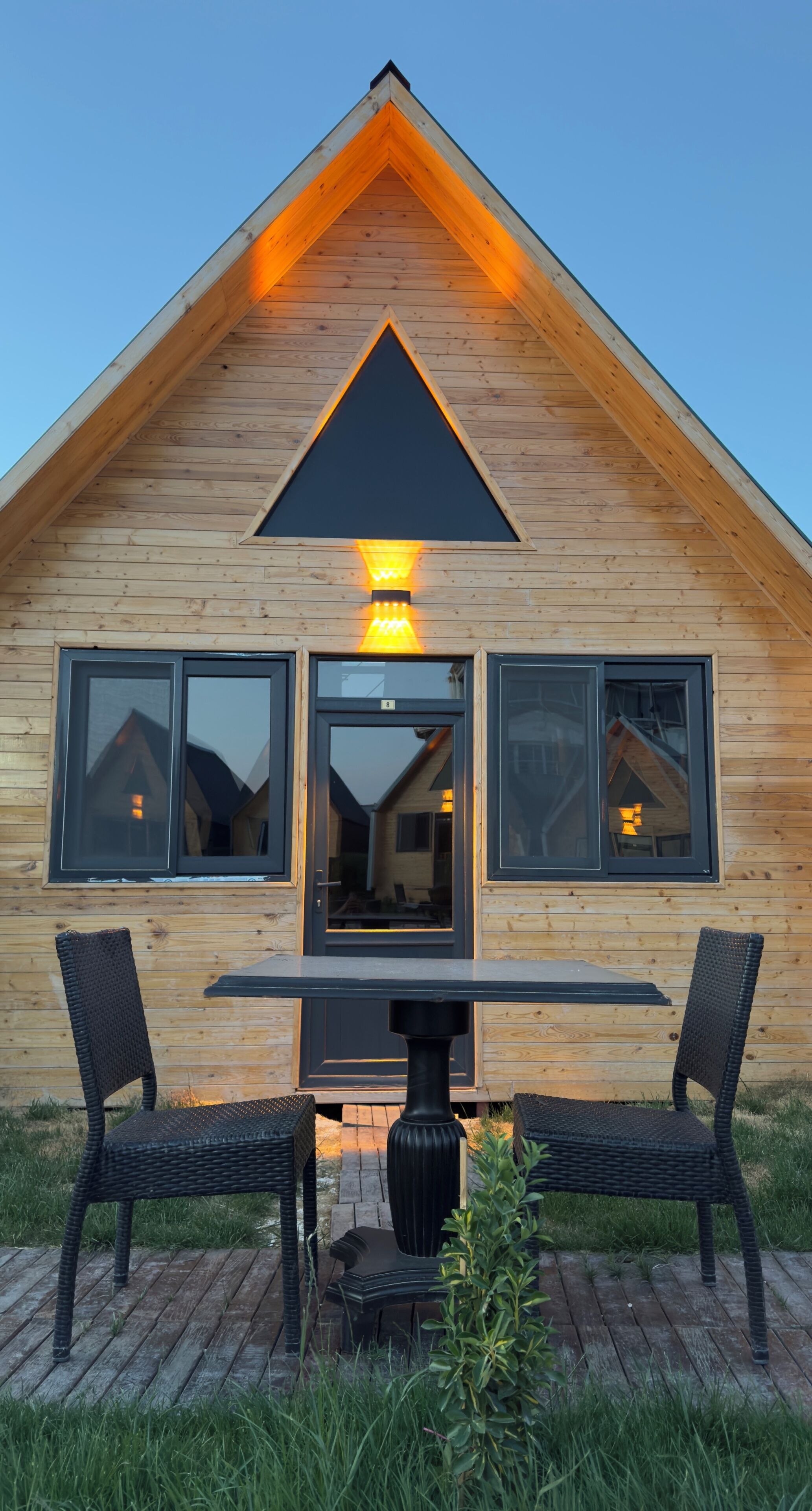 A wooden cabin with a triangular roof, a central table with chairs, and a small garden area.