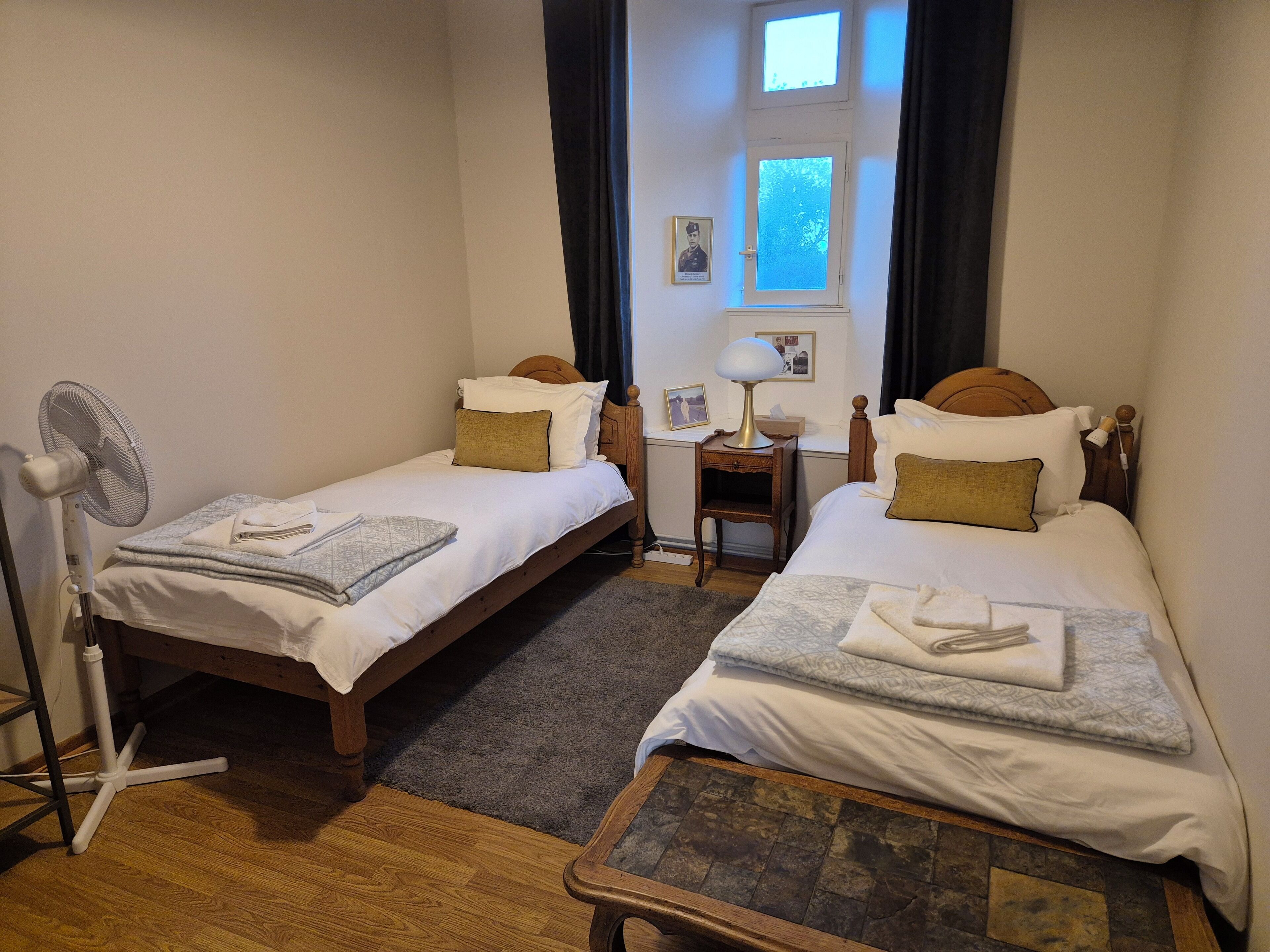 A small, cozy room with two single beds, a desk with a lamp, a chair, and a standing fan.