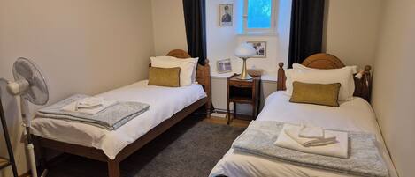A small, cozy room with two single beds, a desk with a lamp, a chair, and a standing fan.