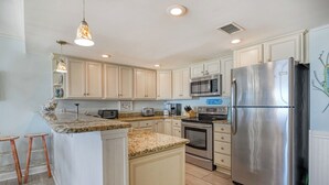 A modern kitchen with light-colored cabinets, a stainless steel refrigerator, a microwave, and an oven. There are stools at the island and a hanging light fixture. - Atalaya Tower 1603 - Three Bedroom, Oceanfront Condo in Garden City Beach (Garden City Beach)