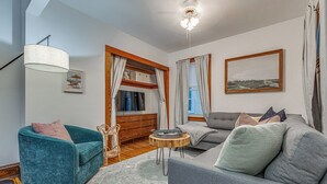 A modern living room with a teal sofa, grey sofa, and a small round table. There is a wooden dresser and a ceiling light fixture. - Fresh Coast Craftsman - Pet Friendly | Near Lake (Sheboygan)