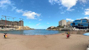 A beach with a sandy shore, a few umbrellas, and a building under construction in the background. - City Centre St. Julian's 1BR Apartment - 4 Guests (Saint Julian's)