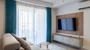 Un saló modern amb un gran televisor de pantalla plana muntat sobre un panell de fusta, un sofà beix amb coixins, una taula rodona petita i una finestra gran amb cortines blaves.