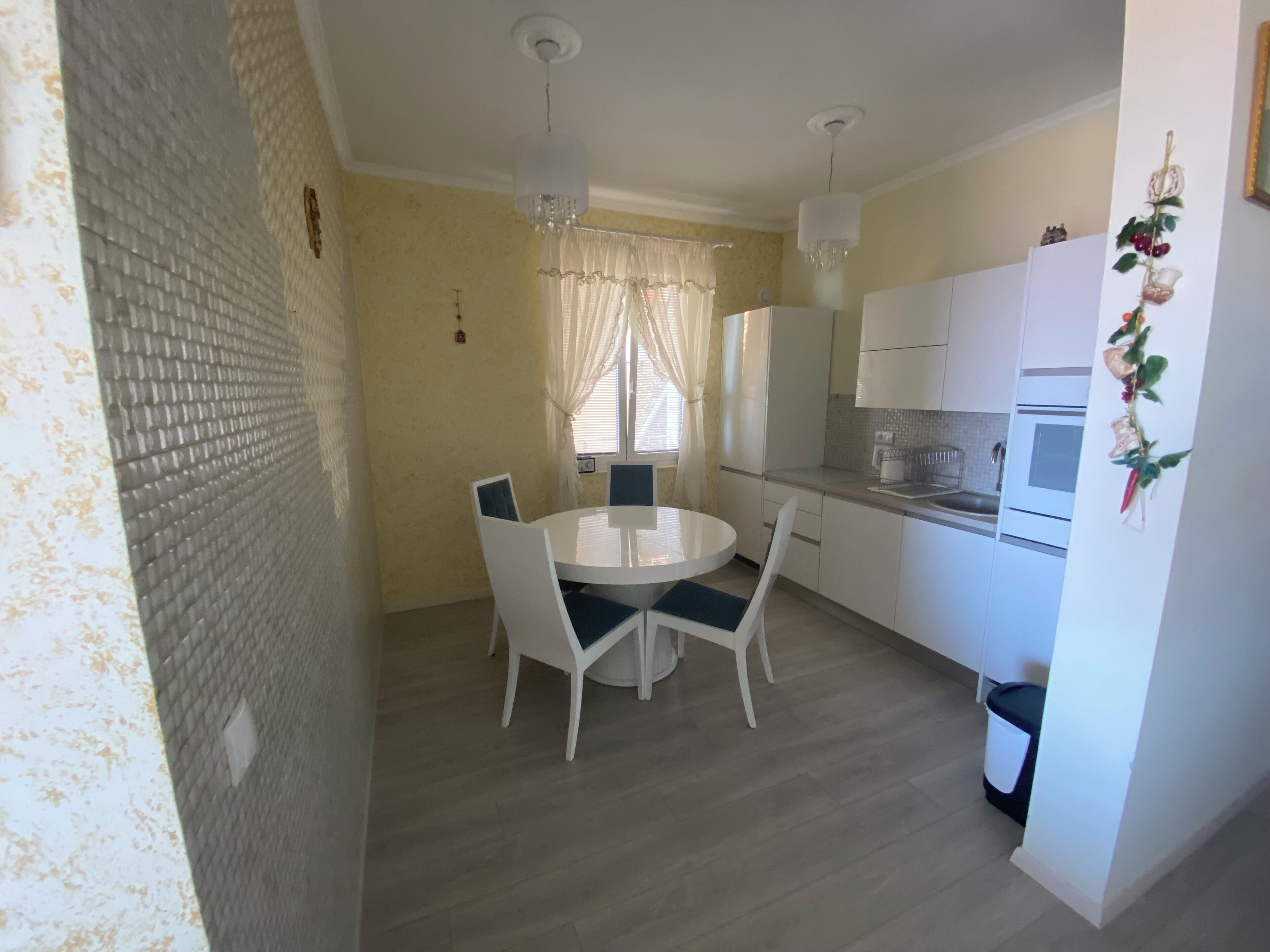A compact kitchen with white cabinets, a round dining table with chairs, and a window with sheer curtains.