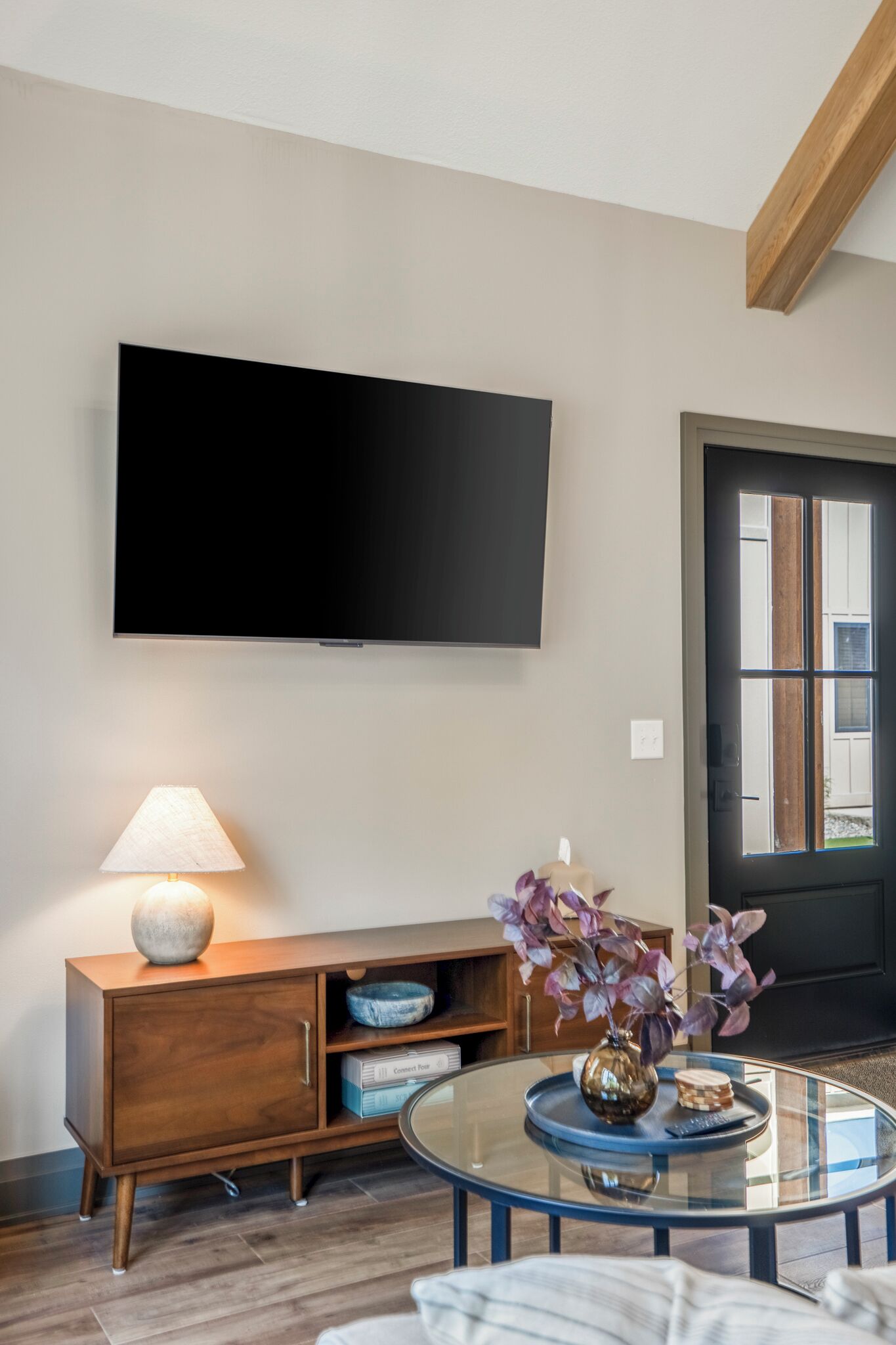 A modern living room with a flat-screen TV mounted on the wall, a wooden cabinet with a lamp, and a glass coffee table with decorative items.