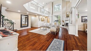 A spacious living area with a high ceiling, wooden flooring, and a large window providing natural light.