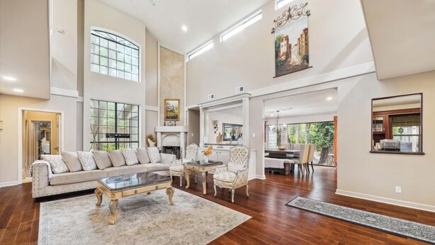 A spacious living room with a large sofa, a coffee table, and a fireplace. There is a dining area with a table and chairs, and a kitchen visible through an open doorway.