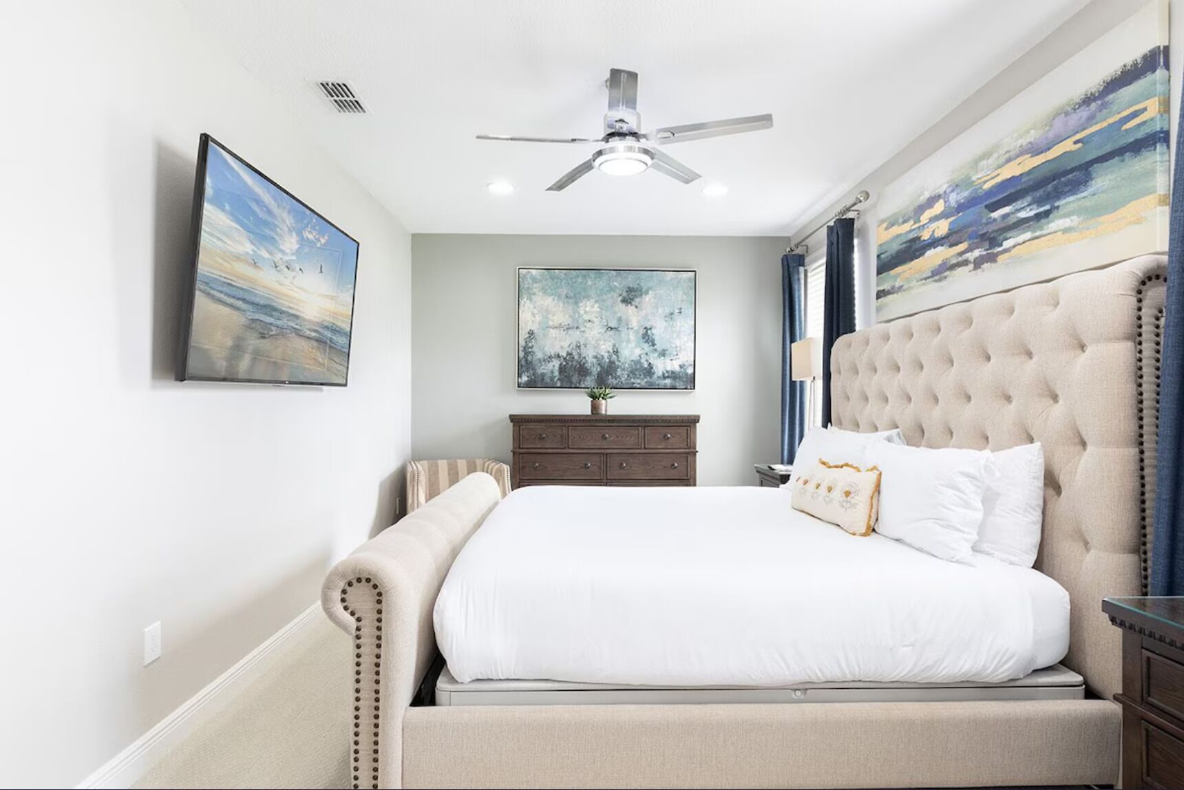 A bedroom with a large bed, a ceiling fan, a dresser, and two paintings on the walls.