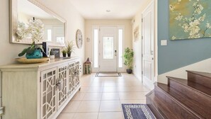 A hallway with a white door, a blue wall with a floral painting, a cabinet with decorative items, and a patterned rug. - Luxury 6BR • Heated Pool Included • Theater •Disney • H2O Perks • No Resort Fees (Kissimmee)