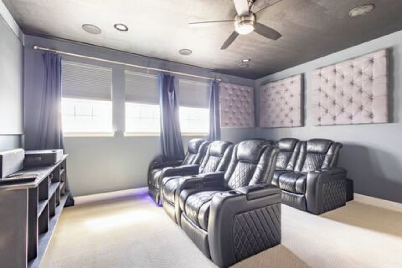 Une salle de cinéma maison équipée d’un ventilateur de plafond, de deux grandes fenêtres avec rideaux et d’un système audio fixé au mur.