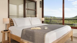 A bedroom with a large bed, two bedside lamps, a bedside table, a window with a view of the outdoors, and a decorative wall piece. - Vista Popoyo 3BR Coastal Retreat with Surf & Mangrove Views (Las Salinas)