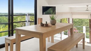 A modern room with a wooden table and bench, a TV mounted on the wall, and large windows offering a view of greenery. - Vista Popoyo 3BR Coastal Retreat with Surf & Mangrove Views (Las Salinas)