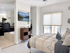 A bedroom with a bed, a TV mounted on the wall, a window with blinds, and a bathroom with a mirror and cabinets.