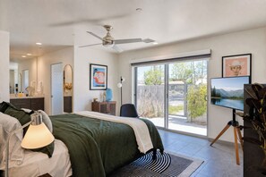 A modern bedroom with a large bed, a ceiling fan, a TV on a tripod, and a sliding glass door leading to a balcony.