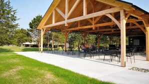 Een houten paviljoen met een eenvoudig, open ontwerp, met een betonnen vloer en metalen stoelen.